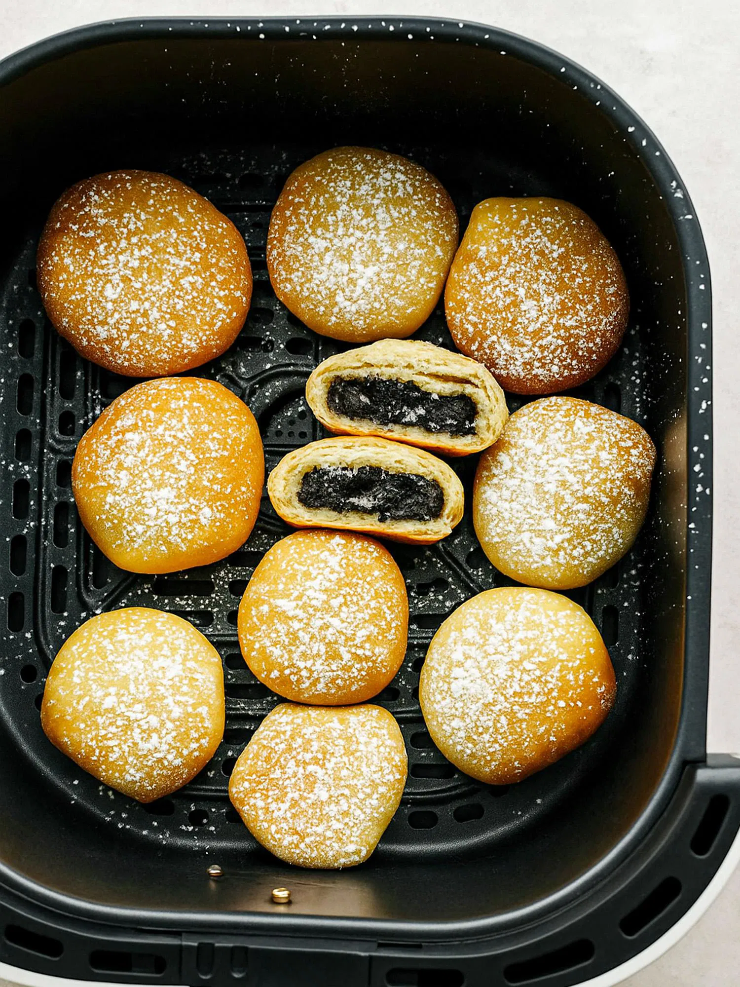 Golden-brown pastries dusted with powdered sugar in an air fryer basket, with one cut open to reveal dark black sesame filling