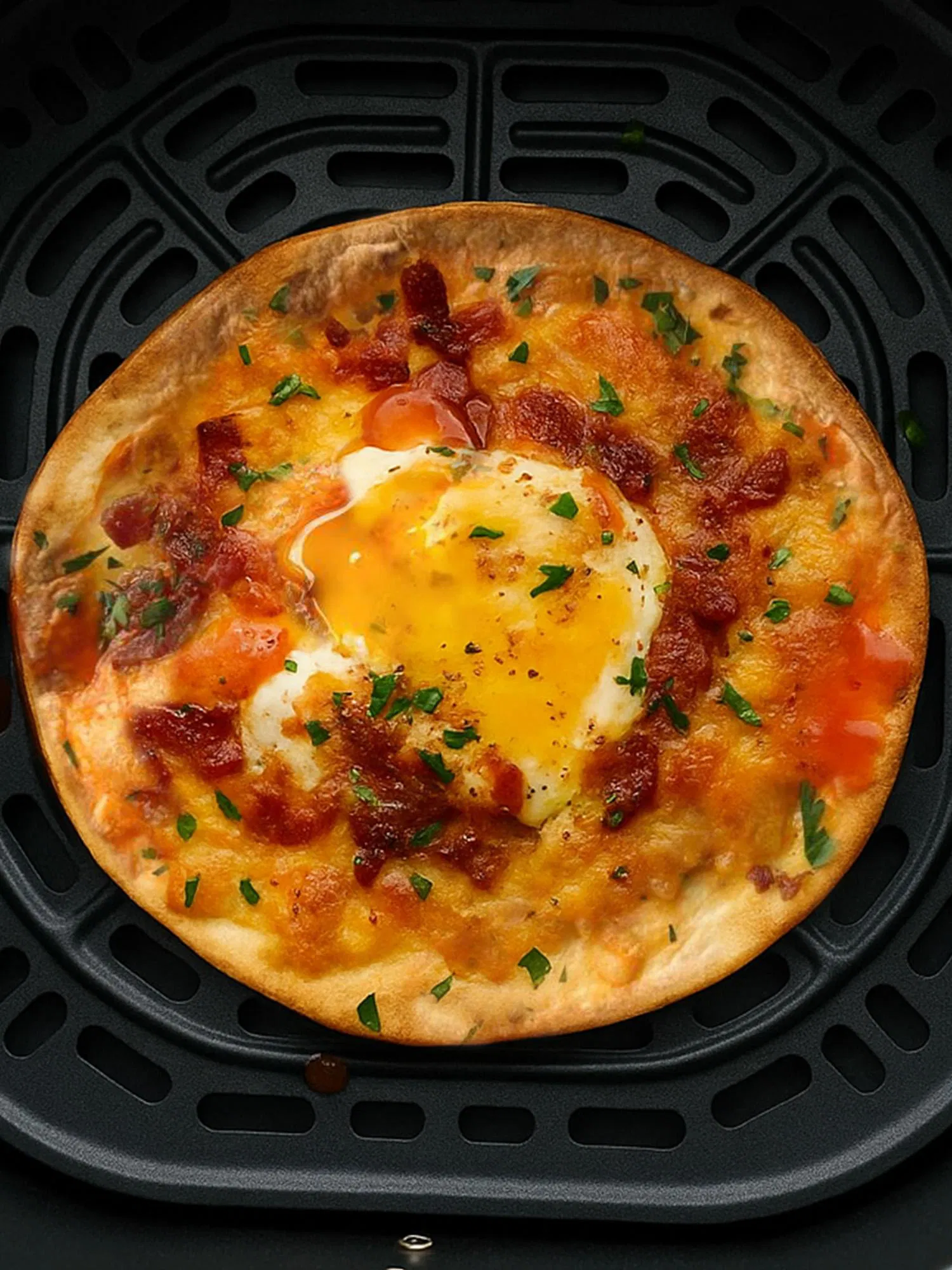 Golden breakfast pizza topped with cheese, bacon, and a sunny-side-up egg sitting in a black air fryer basket