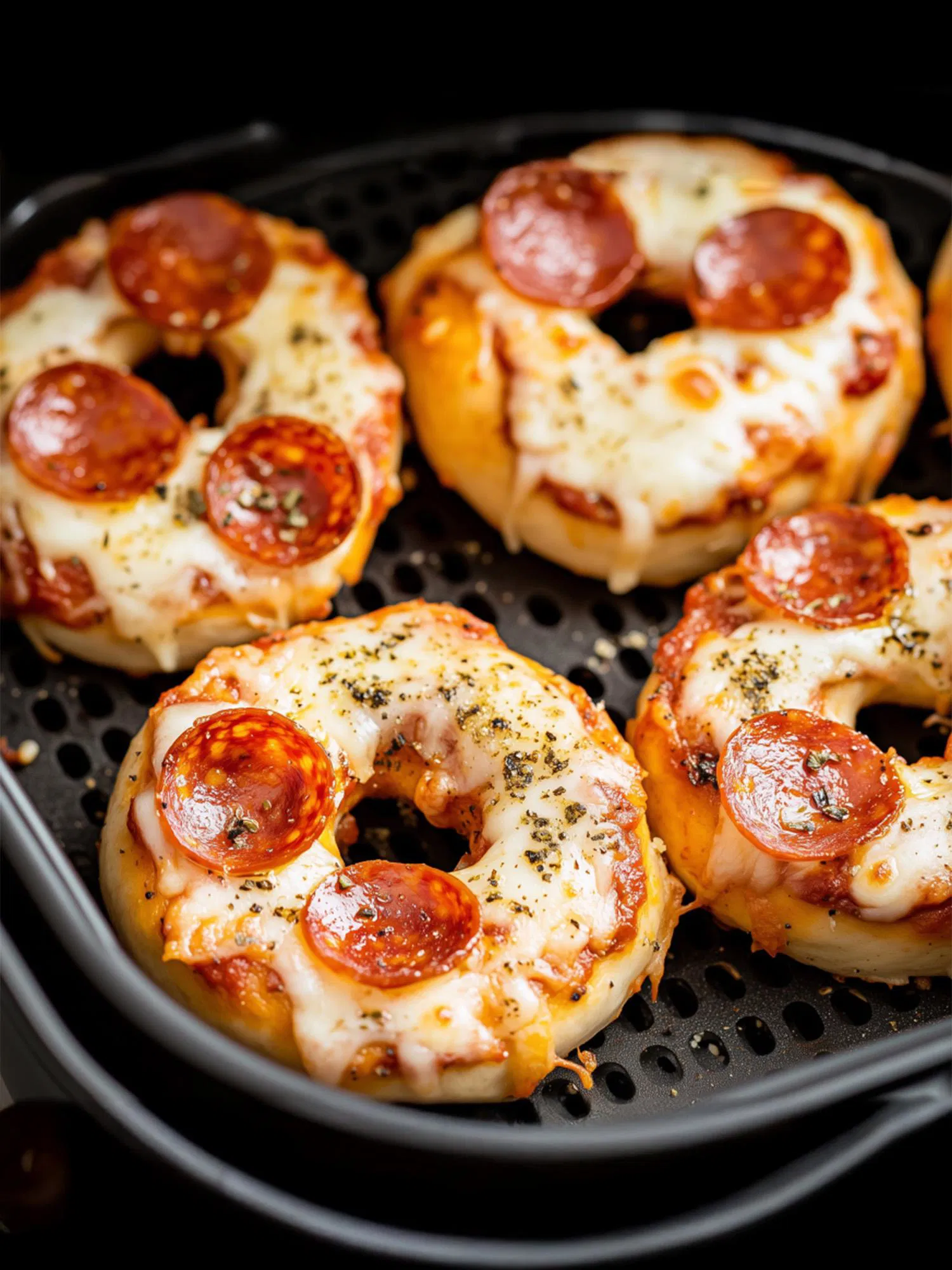 Four pepperoni bagel pizzas with melted cheese and herbs sitting in a black air fryer basket