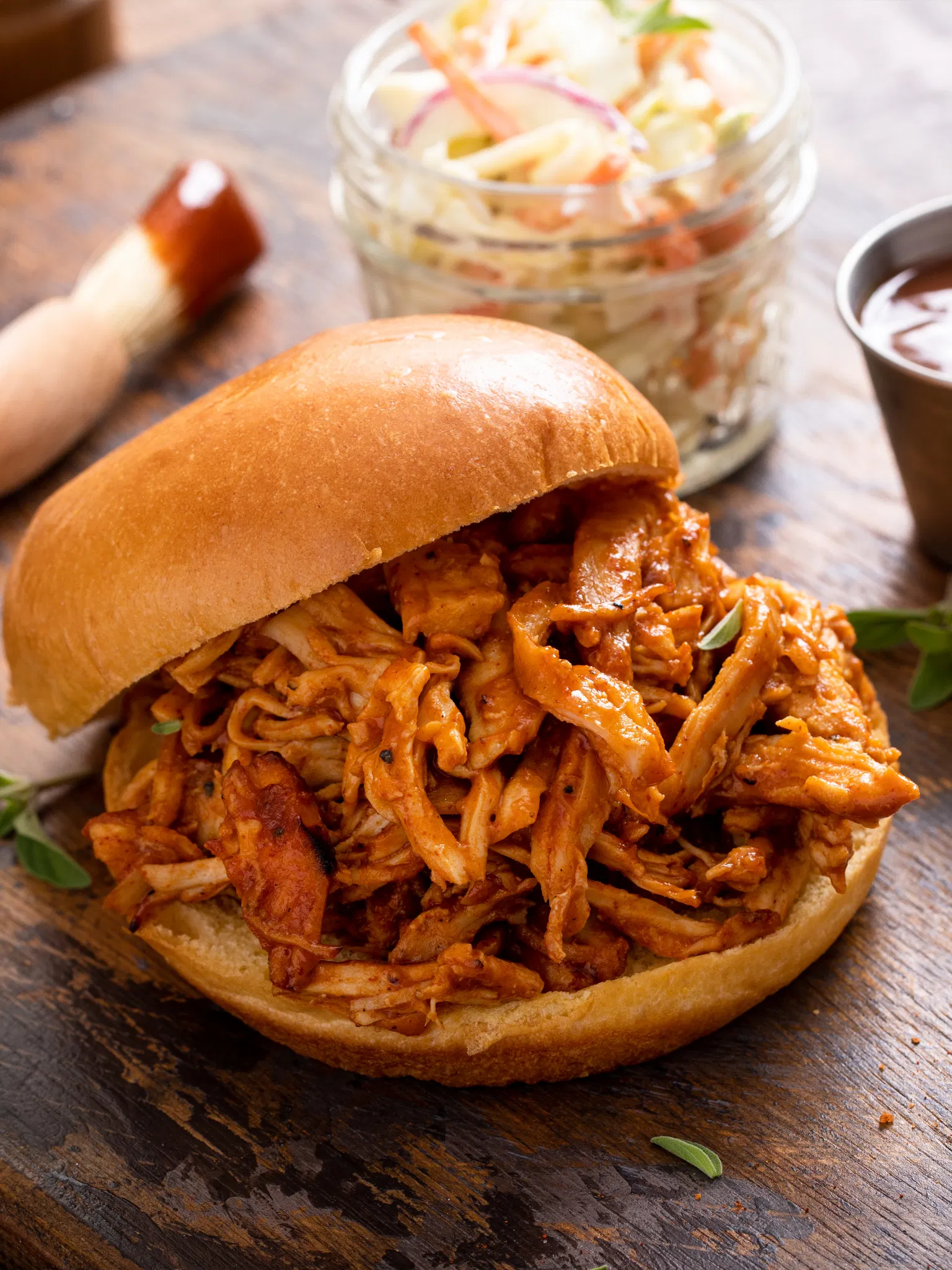 Pulled chicken sandwich with red sauce on a bun, coleslaw in a jar and basting brush on a wooden table