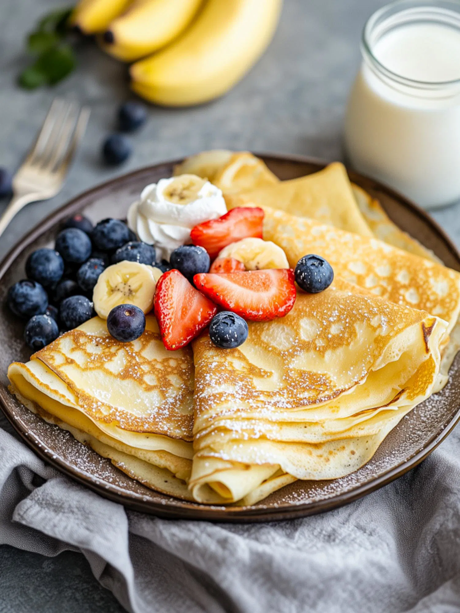 Folded crepes on a brown plate topped with powdered sugar, whipped cream, strawberries, bananas, and blueberries