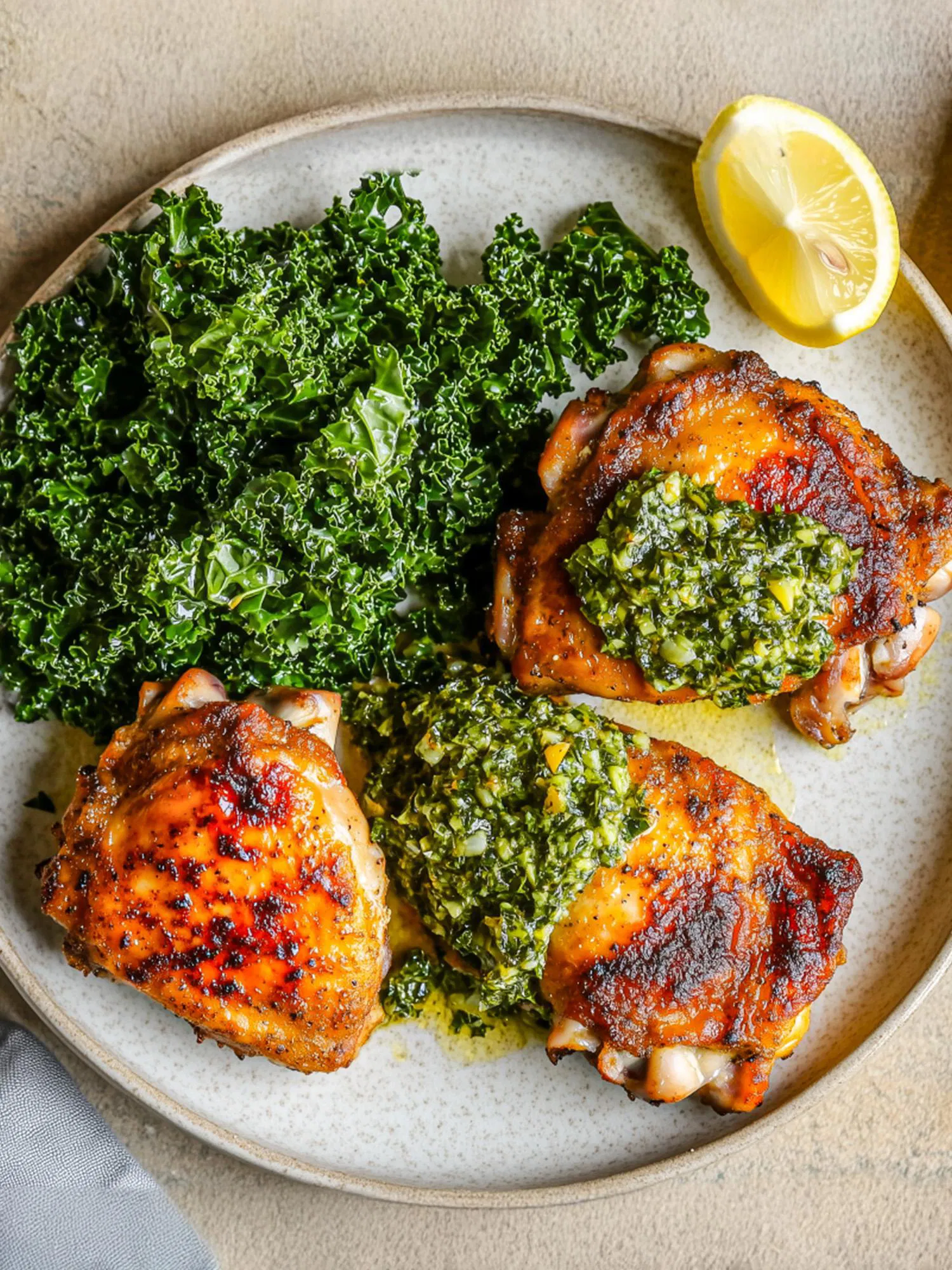 Roasted chicken thighs topped with green chimichurri sauce, served with sautéed kale and a lemon wedge on a speckled ceramic plate.