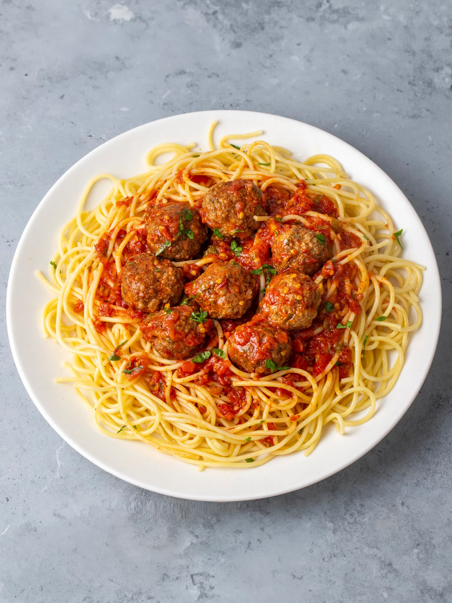 A white plate of spaghetti topped with meatballs in rich red tomato sauce, garnished with fresh parsley on a gray surface.