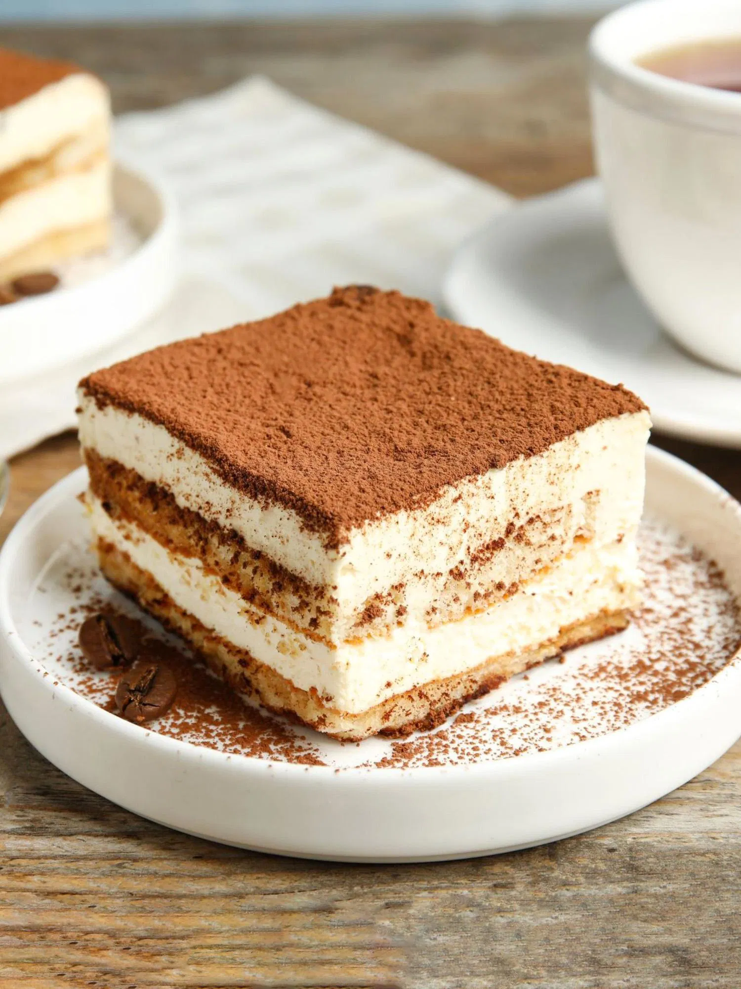 Square slice of classic tiramisu on a white plate with cocoa powder dusting and coffee beans