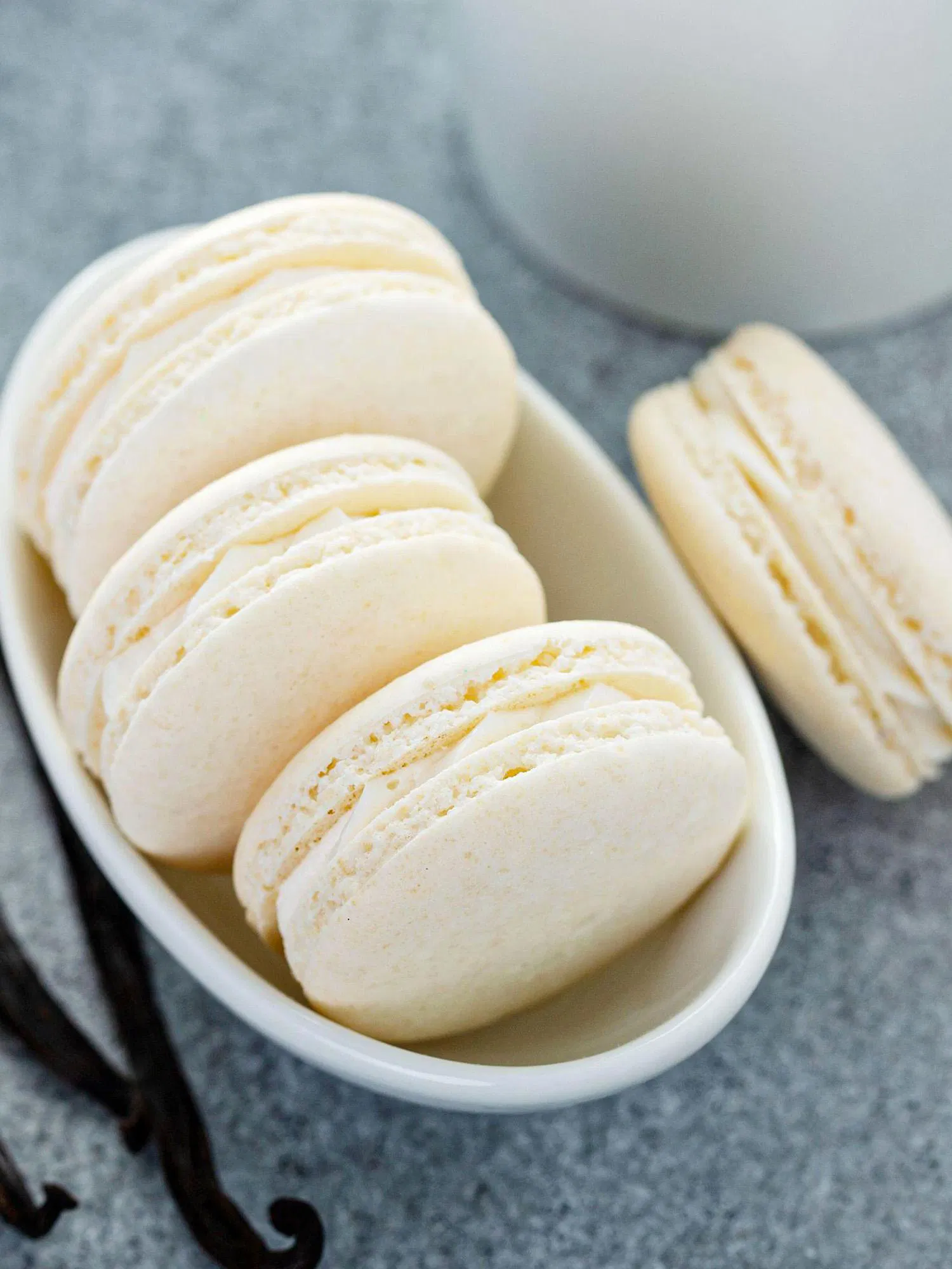 Creamy white vanilla bean macarons lined up in a small white oval dish with whole vanilla beans on a grey surface