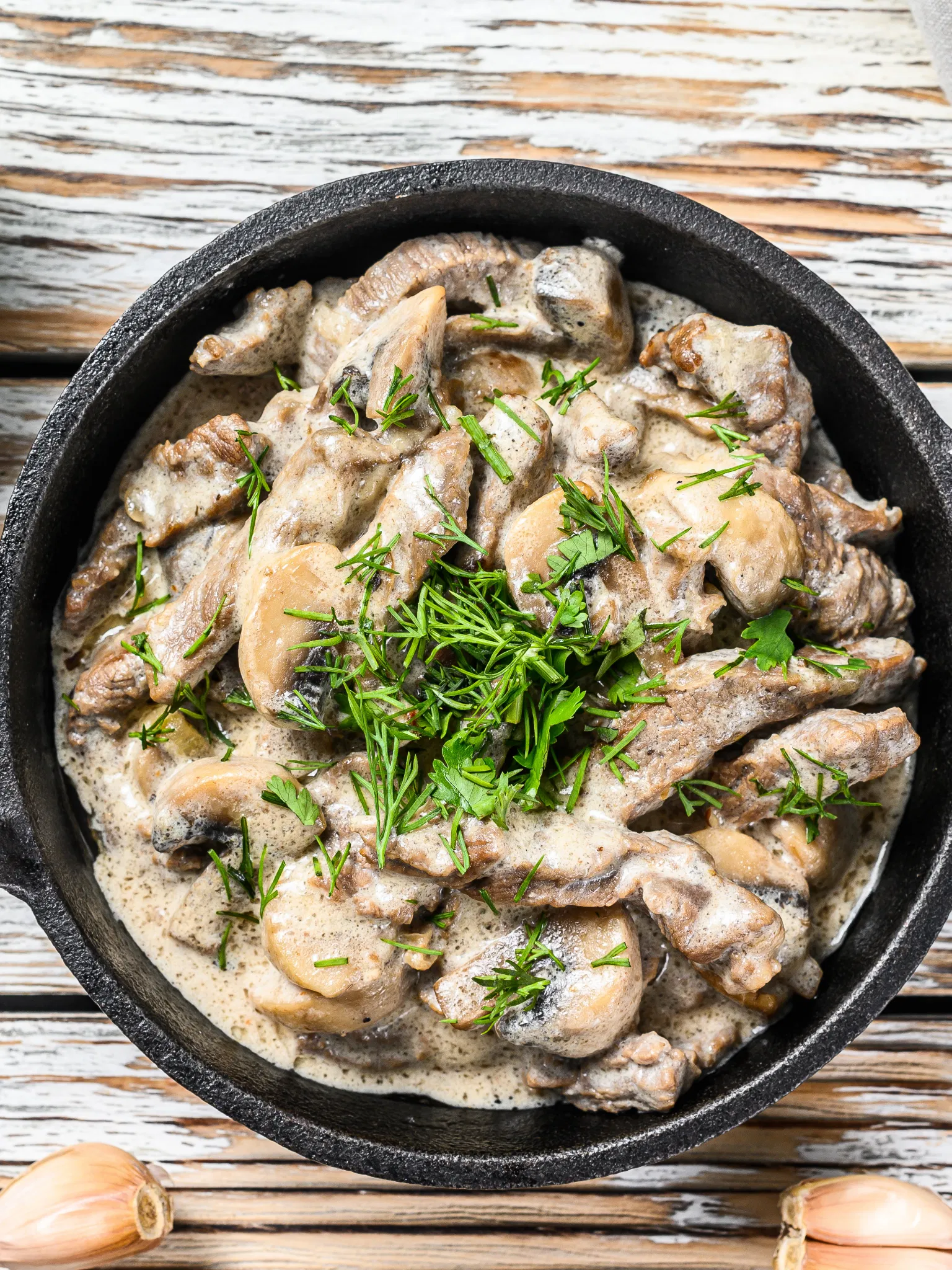 Sliced beef and mushrooms in a creamy sauce served in a black cast iron skillet, garnished with fresh dill and parsley.