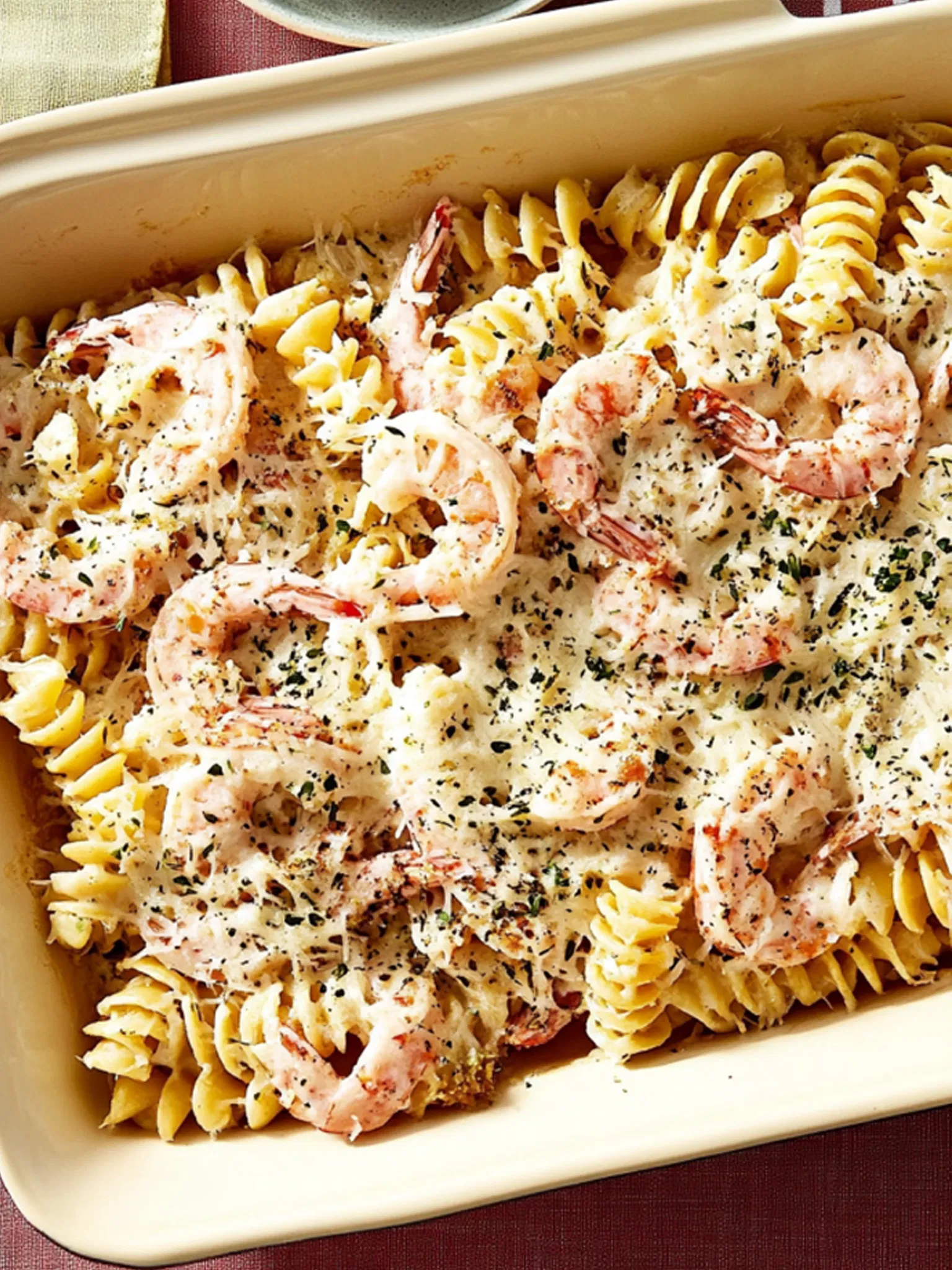 Creamy shrimp and fusilli pasta bake in a beige casserole dish topped with melted cheese and herbs