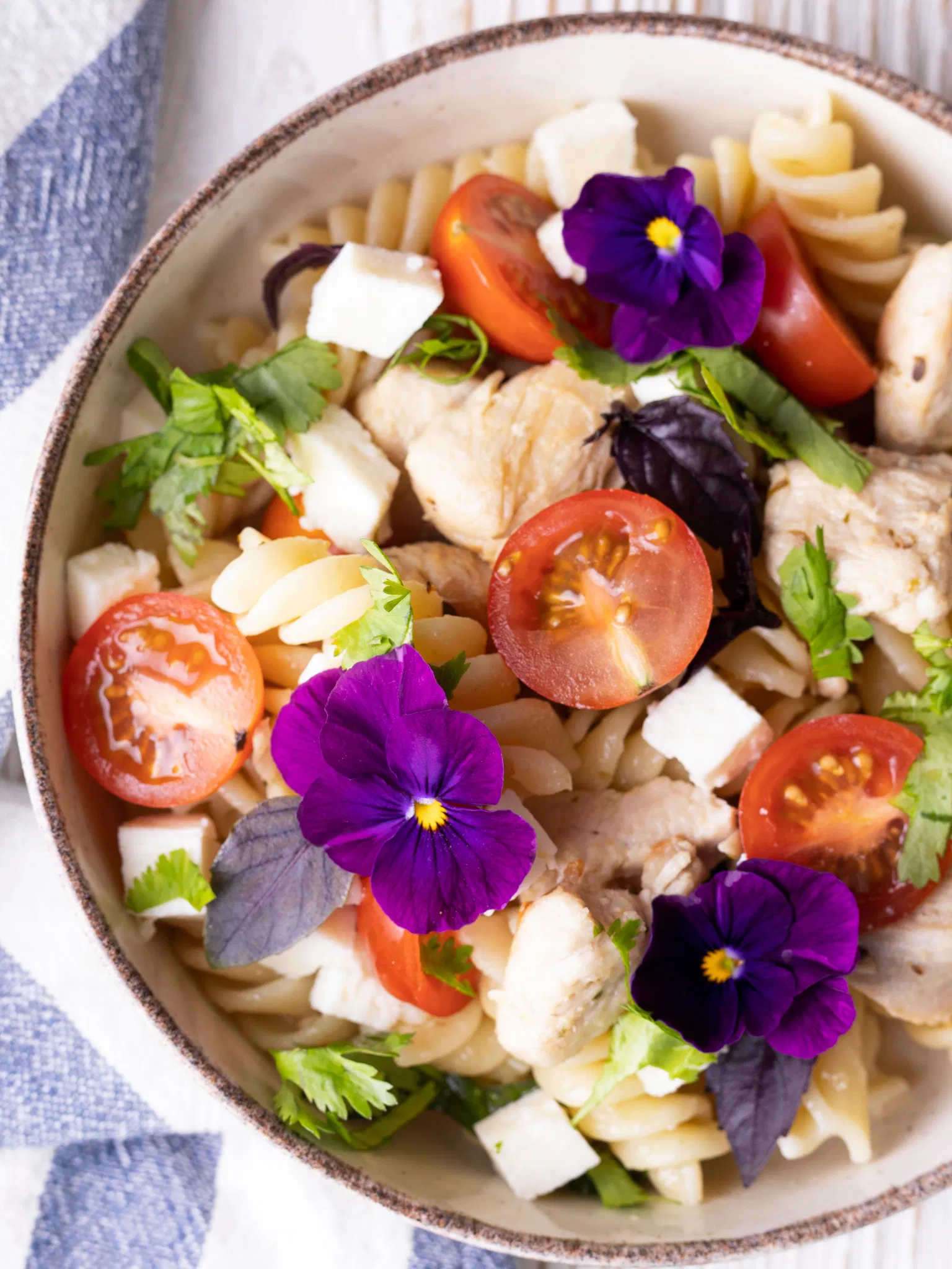 Fusilli pasta salad with chicken, cherry tomatoes, feta cheese cubes, and purple edible flowers in a rustic bowl