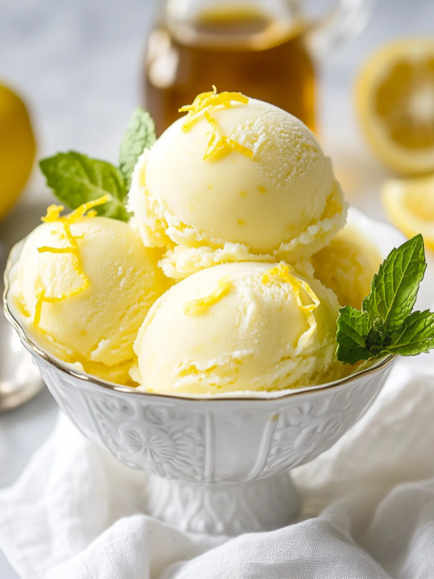 Three scoops of creamy pale yellow lemon ice cream in an ornate white bowl, garnished with fresh mint leaves and lemon zest
