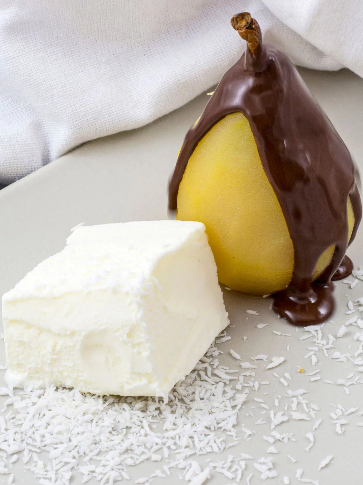 Poached pear drizzled with dark chocolate next to a slice of white coconut cake on a plate with shredded coconut