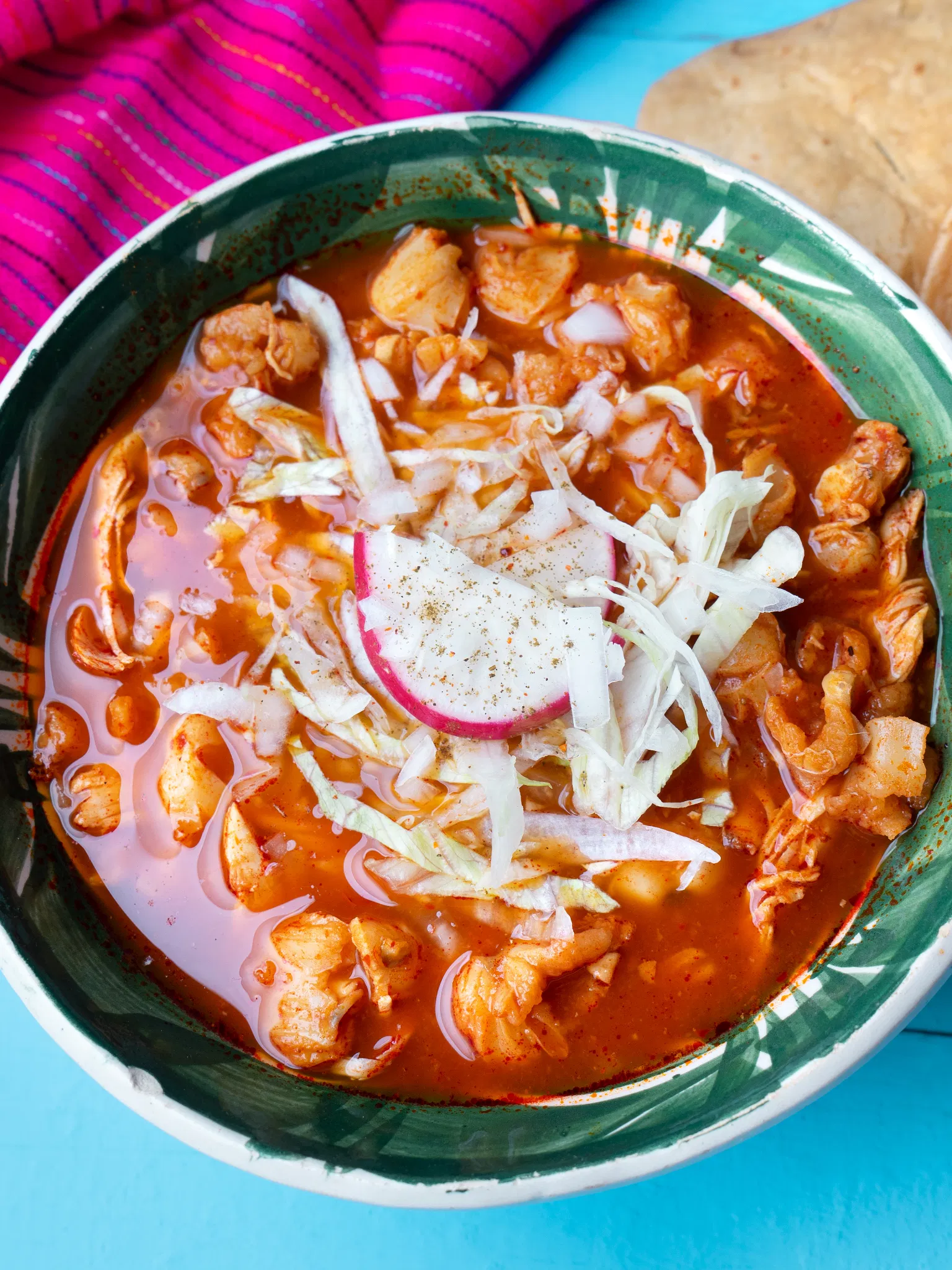 Close-up of a green bowl filled with red soup, shredded chicken, cabbage, and a slice of radish