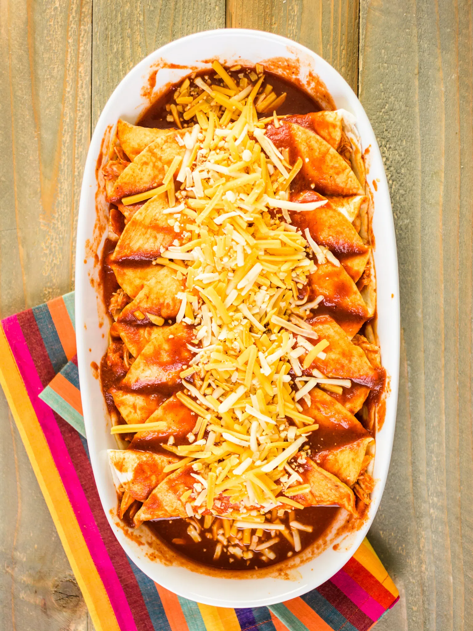 White oval dish filled with rolled tortillas covered in red sauce and shredded cheese on a wooden table