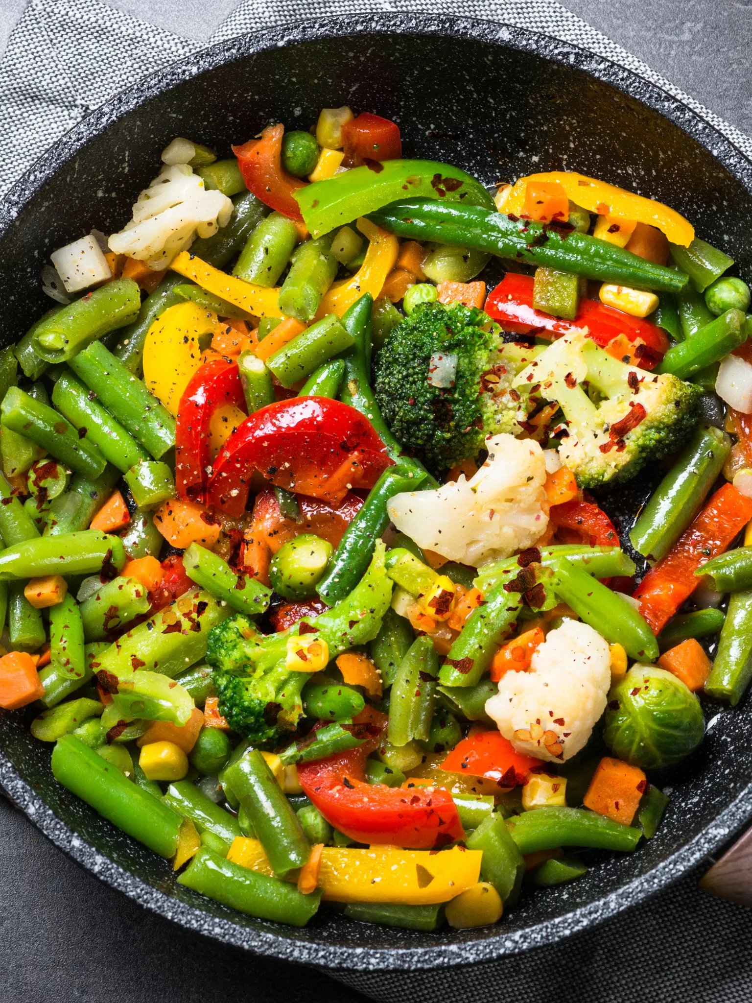 Colorful mixed vegetables like green beans, broccoli, peppers, and cauliflower sautéed in a black pan with red pepper flakes
